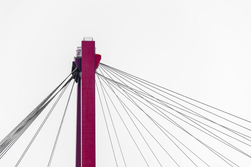 Afbeelding: Een strakke, felrode brugpijler die verticaal uit de onderkant van het beeld omhoog schiet tegen een witte lucht, met meerdere kabels die naar rechts weglopen