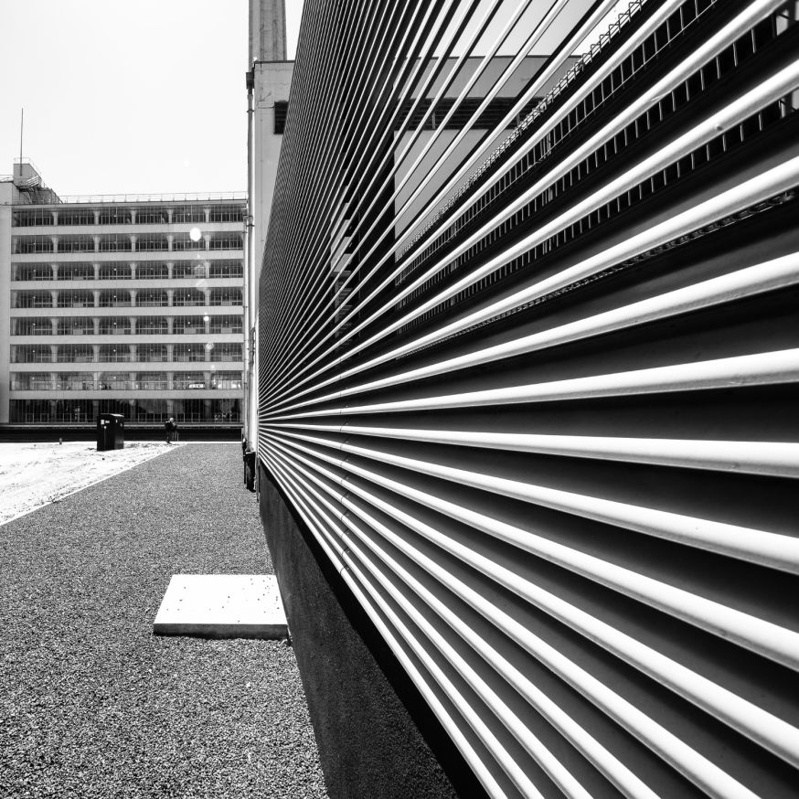 Afbeelding: Een zwart-wit foto van een gevel met horizontale lamellen op de voorgrond. Op de achtergrond is een groot gebouw met een strak grid van ramen zichtbaar