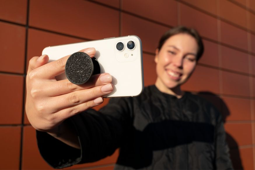 Afbeelding: Een vrouw die een selfie maakt met een witte smartphone waarop een ronde, zwarte glinstering-grip (pop-grip) is bevestigd
