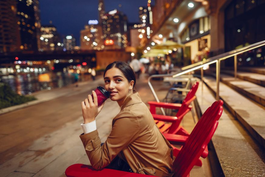 Afbeelding: Jonge vrouw drinkt koffie langs de waterkant in een sfeervol verlichte stad bij avond