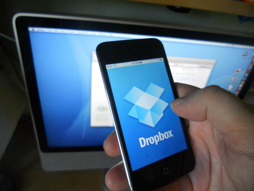 Afbeelding: Een hand houdt een iPod Touch vast waarop het blauwe Dropbox-logo op het scherm staat, met op de achtergrond een iMac-computer